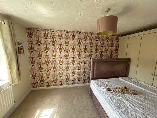 A bedroom with patterned wallpaper, a beige carpet, and a bed with an unmade mattress.